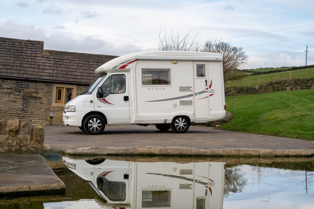 A 2006 Auto-Sleepers Nuevo EK, adorned with red and grey decals, is parked on a driveway beside a house. Its reflection shimmers in the body of water in the foreground. The background features green grass, a stone wall, and a cloudy sky.