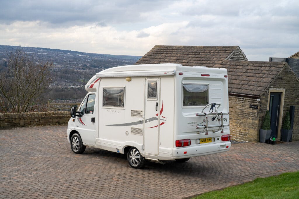 A 2006 Auto-Sleepers Nuevo EK camper van is parked on a paved driveway in front of a stone building with a sloped roof. The van, equipped with a bike rack attached to the rear, is surrounded by a scenic view of a hilly landscape under a cloudy sky.