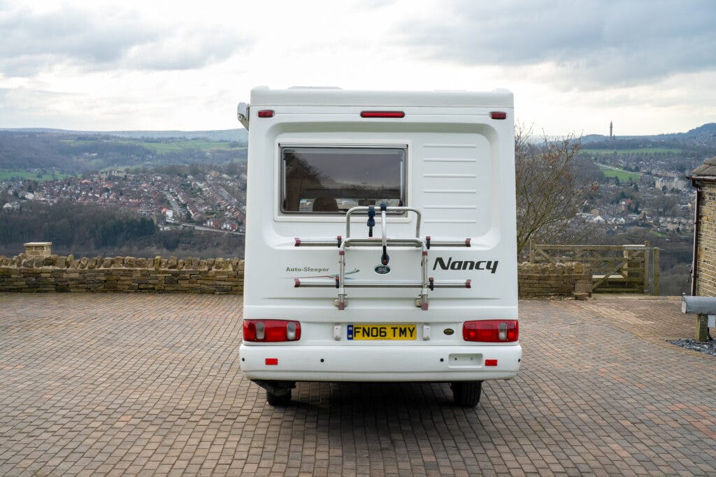 A white 2006 Auto-Sleepers Nuevo EK camper van labeled "Nancy" is parked on a paved surface, offering a scenic view of a town and hills in the background. The van features a rear window, bike rack, and license plate "FN06 TMY." The sky is partly cloudy.