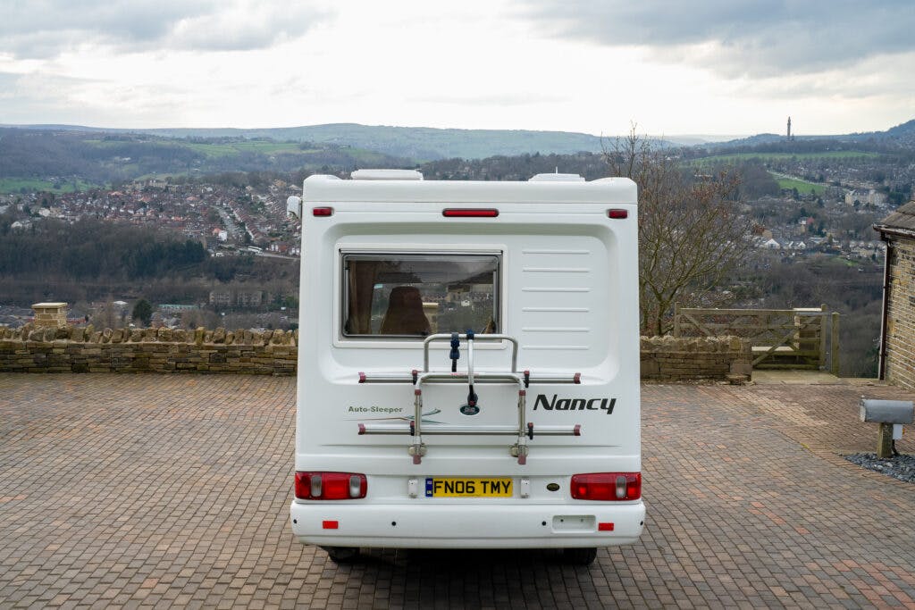 A 2006 Auto-Sleepers Nuevo EK white camper van is parked on a brick driveway overlooking a scenic landscape with rolling hills and a small town in the distance. The rear of the camper van features the word "Nancy" above the license plate.