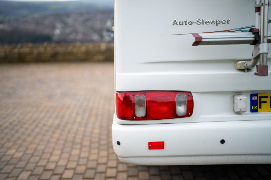 Close-up view of the rear corner of a 2006 Auto-Sleepers Nuevo EK camper van, focusing on the tail light and part of the license plate with visible registration letters 'F'. Background includes a stone wall and an out-of-focus landscape with hills.
