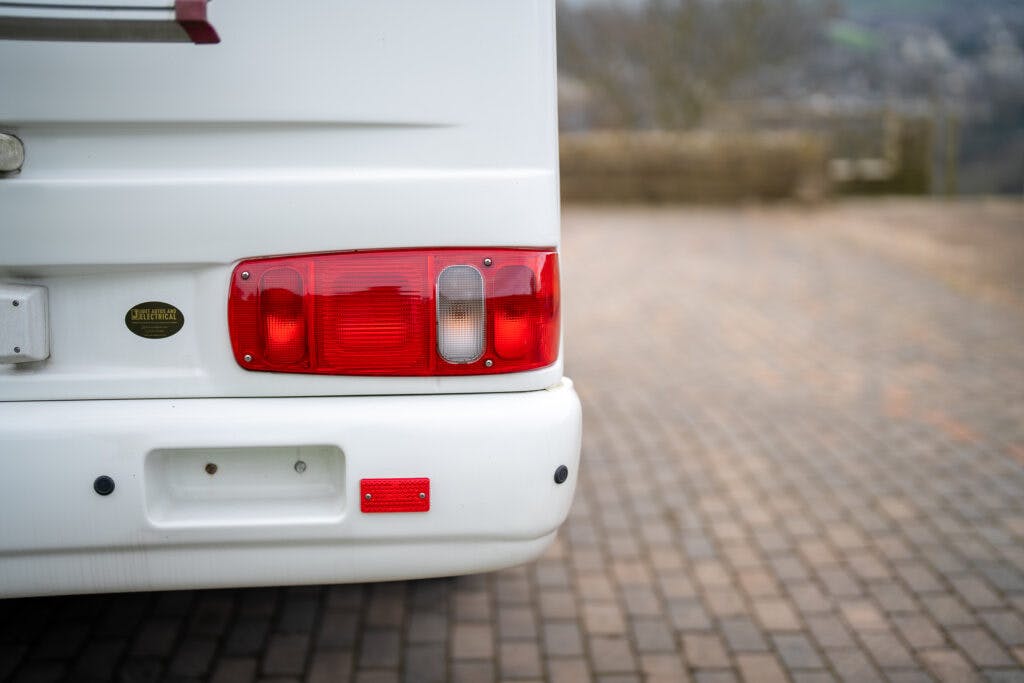 Close-up of the rear left end of a 2006 Auto-Sleepers Nuevo EK camper van showing red and white tail lights on a paved surface. The vehicle has a white bumper with a red reflective sticker. The background is blurred, with a stone wall and some greenery in the distance.