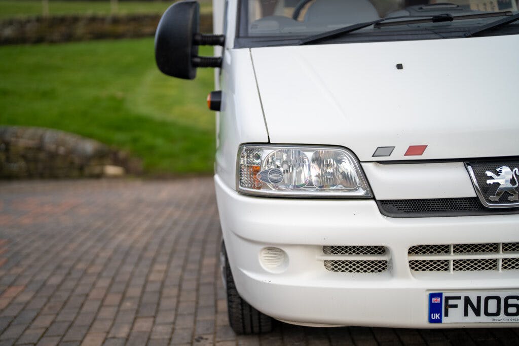 A close-up view of the front of a white Peugeot van, possibly a 2006 Auto-Sleepers Nuevo EK model. The headlight and front grille are visible, along with part of the license plate displaying "FND." The vehicle is parked on a brick driveway, and a grassy area with a stone wall is in the background.