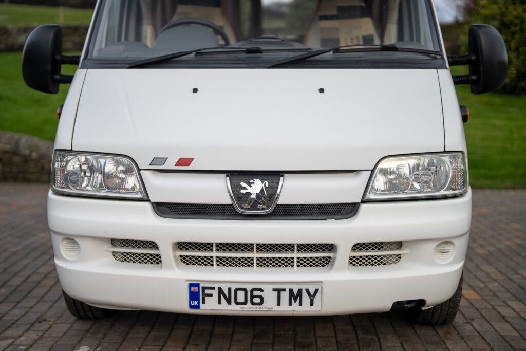 A white Peugeot van with a UK license plate reading "FN06 TMY" is parked on a brick pavement. The 2006 Auto-Sleepers Nuevo EK features the Peugeot logo, two rectangular headlights, and two small red and gray rectangles positioned above the grille.
