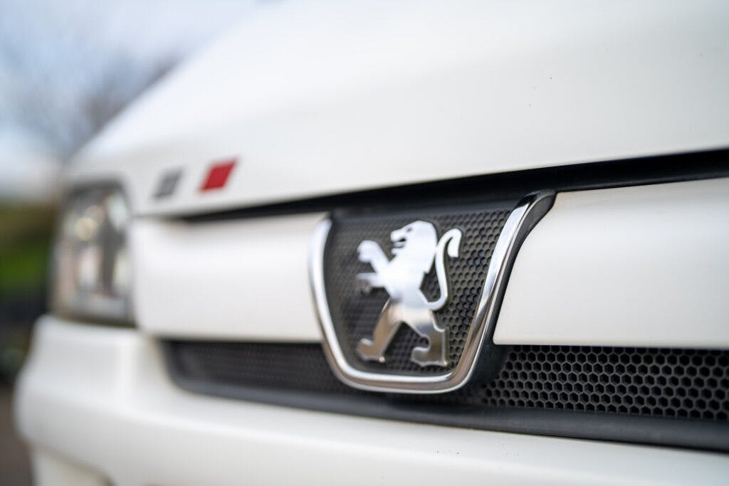 Close-up of the front grille of a white Peugeot car, displaying the iconic Peugeot logo featuring a silver lion on a shiny black background. The out-of-focus headlamp of the 2006 Auto-Sleepers Nuevo EK is visible in the background on the left side.