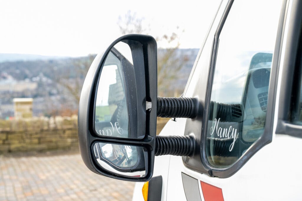 A close-up of the side mirror of a 2006 Auto-Sleepers Nuevo EK, showing reflections of the surrounding outdoor area. The side mirror has two reflective surfaces, and there is a name "Nancy" on the side window of the vehicle. The background features a stone wall and trees.