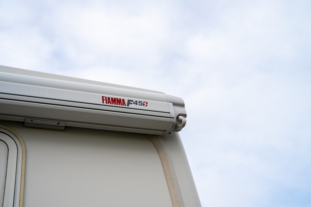 A close-up view of the edge of a 2006 Auto-Sleepers Nuevo EK with a Fiamma F45S awning installed. The awning is white with the brand name and model number in red letters, set against a partly cloudy sky.