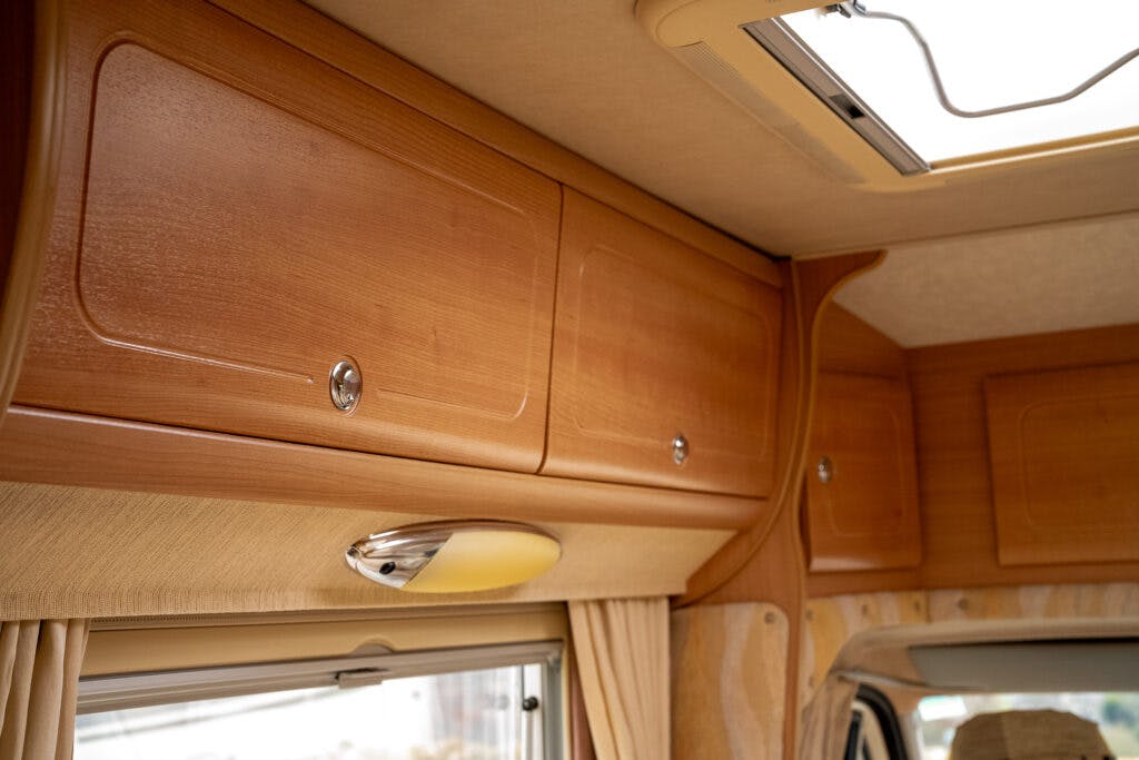 This image shows wooden storage cabinets inside a 2006 Auto-Sleepers Nuevo EK RV. The cabinets have sleek handles and are mounted above a window with beige curtains. An overhead light fixture is visible below the cabinets. The ceiling has a roof vent allowing natural light in.