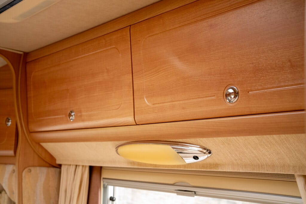 A close-up image of wooden overhead storage cabinets in a 2006 Auto-Sleepers Nuevo EK. The cabinets have a smooth, polished finish and metallic round handles. Below the cabinets, there is a modern oval light fixture with a chrome edge and frosted glass center.
