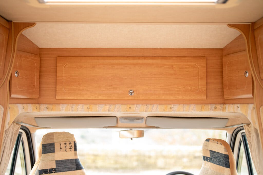 The image showcases the front interior of a 2006 Auto-Sleepers Nuevo EK camper van. A wooden storage cabinet sits above the windshield, while two front seats with fabric covers provide comfort. The ceiling light is also visible.