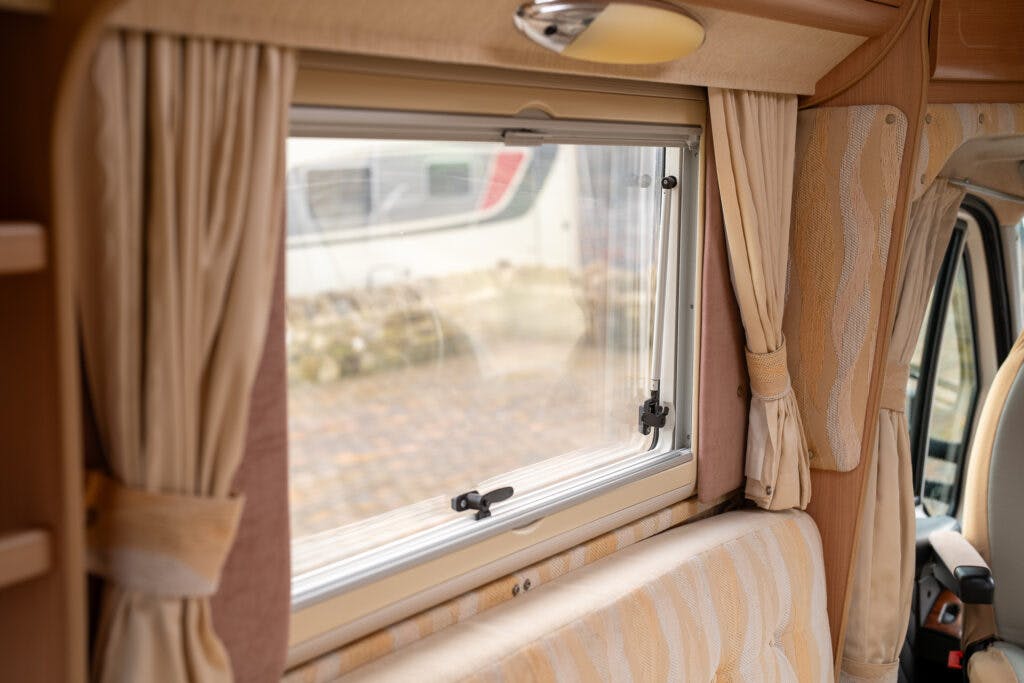 The image shows the interior of a 2006 Auto-Sleepers Nuevo EK, focusing on a window with beige curtains tied back. Below the window is a cushioned seat with matching beige upholstery. The window is partially open, and an exterior view of another vehicle is visible.