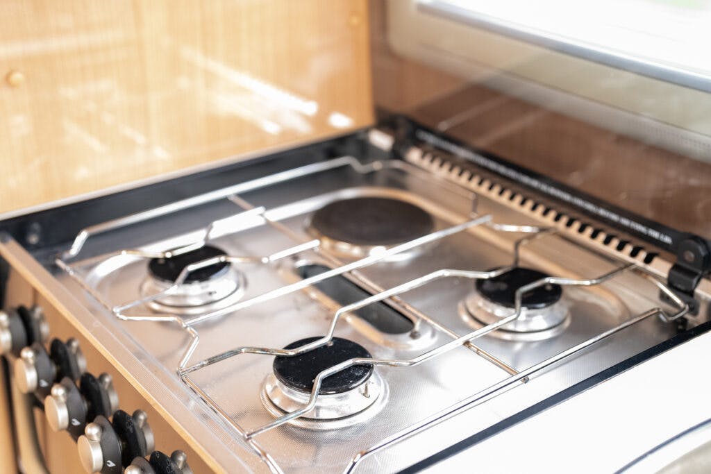 A close-up of a stainless steel gas stove with four burners and black knobs on the left side, reminiscent of the 2006 Auto-Sleepers Nuevo EK. A metal grate covers the burners, and there is a reflective glass panel in the background. The stove is installed in a light-colored wooden cabinet.