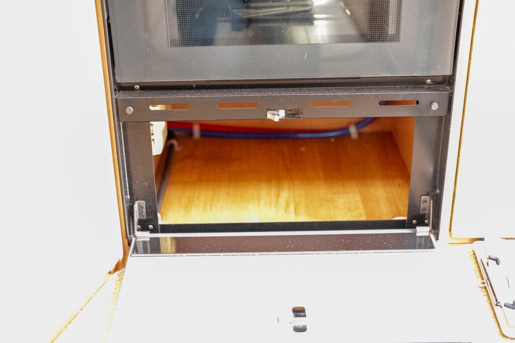 An open oven door reveals the interior of the appliance with an empty cooking chamber. The metallic frame and wooden kitchen floor beneath are visible, surrounded by an orange-brown cabinet. Blue and red cables lie on the floor behind it, reminiscent of a well-maintained 2006 Auto-Sleepers Nuevo EK’s electrical setup.