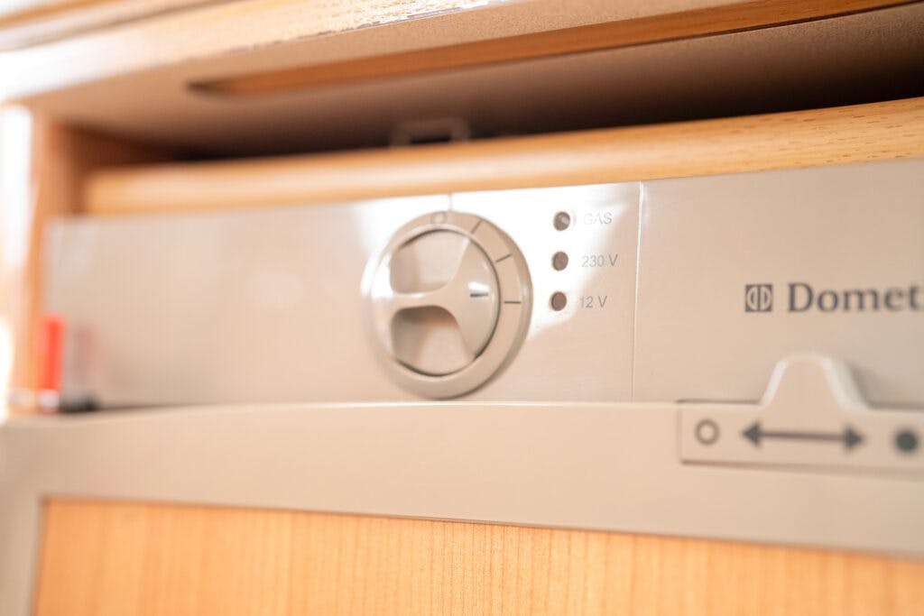 Close-up of a control panel on a Dometic appliance in the 2006 Auto-Sleepers Nuevo EK, featuring a round knob and indicator lights for gas, 230V, and 12V power options. The panel is set in a wooden housing.