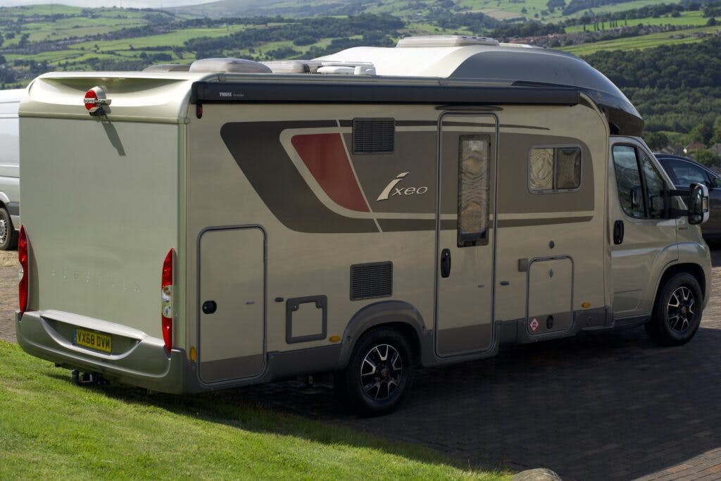 A grey 2018 Burstner Ixeo TL680 G motorhome with a silver trim and maroon accent is parked on a paved surface overlooking a green, hilly landscape. The motorhome has its windows closed and features a bike rack at the rear. The vehicle bears a license plate reading "KX68 DNV.