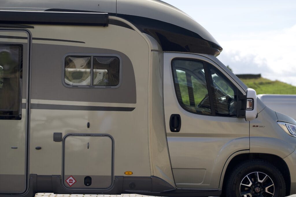 Side view of a 2018 Burstner Ixeo TL680 G motorhome with black accents. The image shows part of the cabin, including a side mirror, windows, and a door. The sky is partly cloudy, and some greenery is visible in the background.