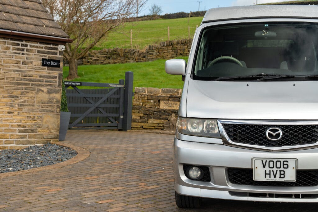 A silver 2006 Mazda Bongo Friendee van is parked on a brick driveway near a stone building with a sign "The Barn." The driveway leads to a wooden gate, behind which are grassy fields and trees. A stone retaining wall is visible in the background. The van's license plate reads "VO06 HVB.