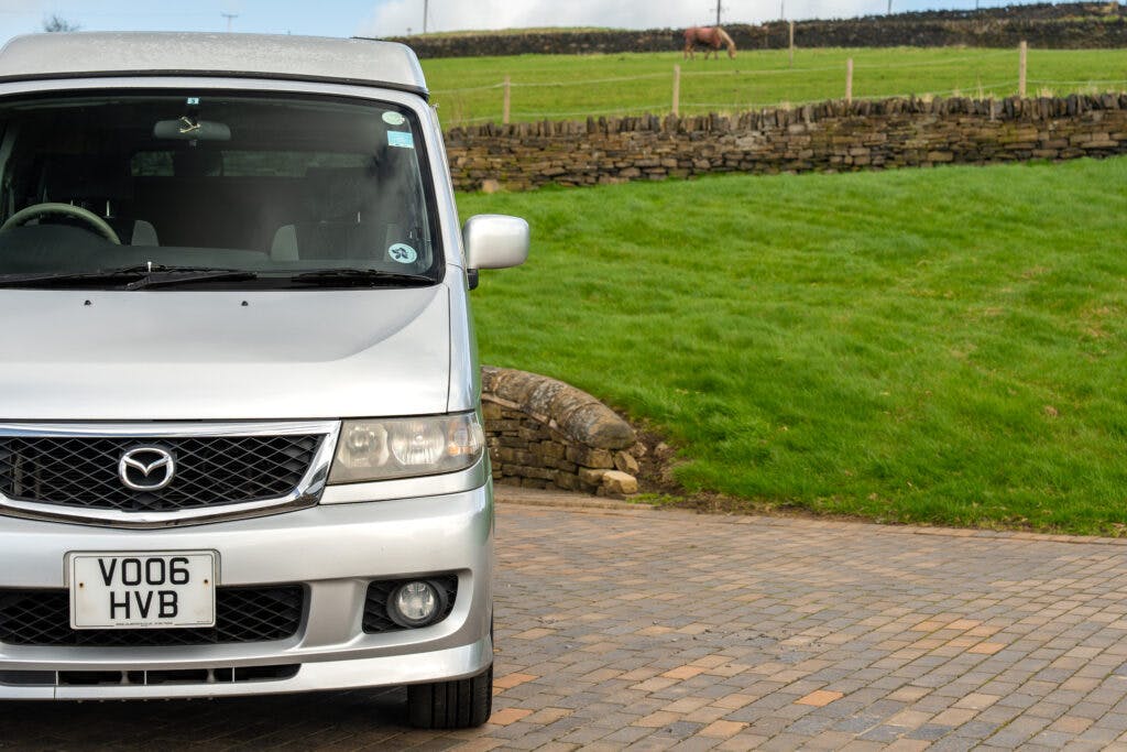 A partial view of a silver 2006 Mazda Bongo Friendee with the license plate "VO06 HVB" parked on a paved surface. In the background, there is a grassy area with a stone wall and a horse grazing.