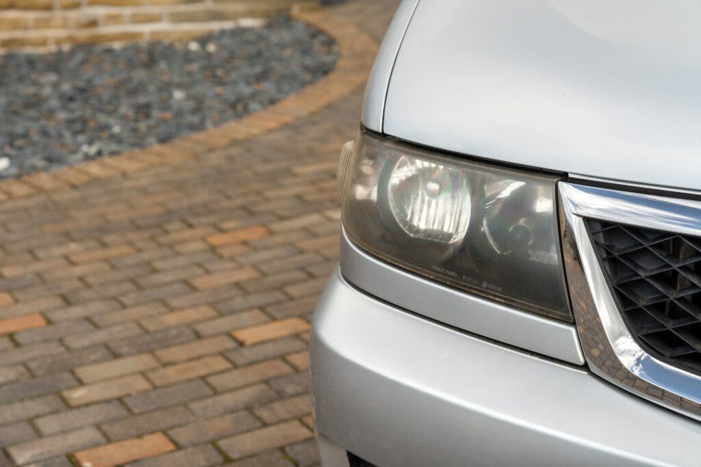 Close-up of a silver 2006 Mazda Bongo Friendee's headlight and part of the front bumper. The car is parked on a paved driveway with a rock garden area in the background. The image focuses on the vehicle’s left headlight and part of the grille.