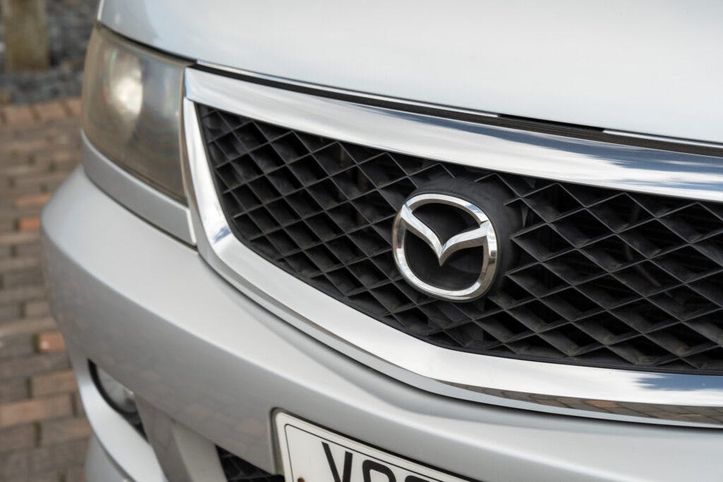 Close-up of the front grille of a 2006 Mazda Bongo Friendee, featuring the Mazda logo in the center. The car is silver with a prominent chrome accent around the grille. The image also shows a portion of a license plate at the bottom.