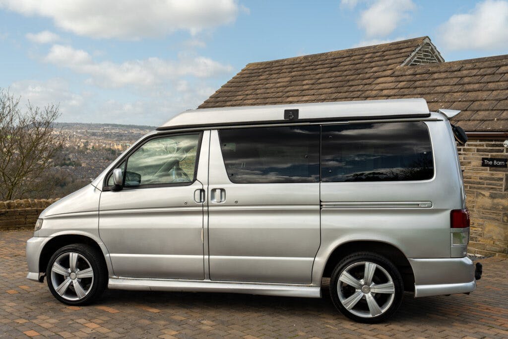 A 2006 Mazda Bongo Friendee silver camper van is parked on a brick-paved surface. It has a raised roof, tinted windows, and alloy wheels. A stone building with a tiled roof is in the background, and a distant hilly landscape can be seen under a partly cloudy sky.