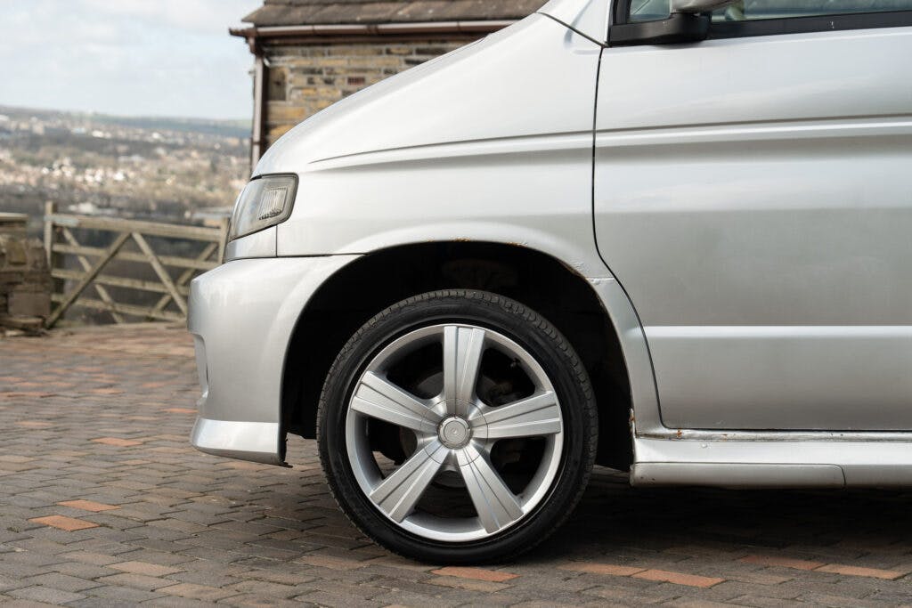 A close-up of a 2006 Mazda Bongo Friendee's front left wheel is shown. The van has shiny alloy wheels with five spokes and is parked on a cobblestone surface. The image reveals a portion of the van's front, featuring slightly worn paintwork near the wheel arch.
