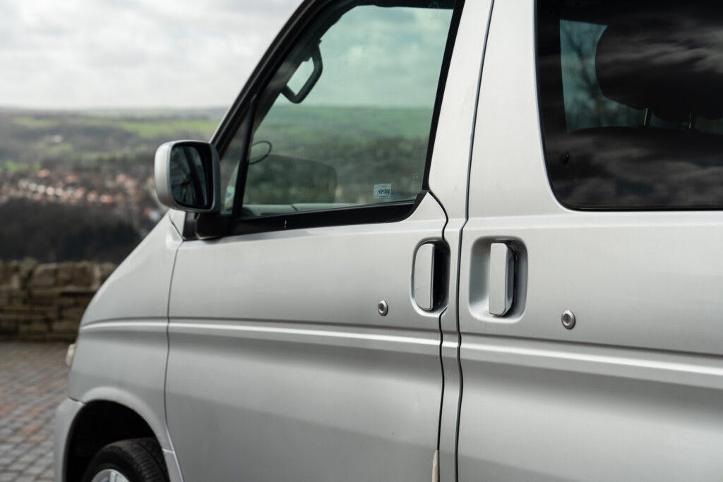 A close-up of a 2006 Mazda Bongo Friendee's side door featuring a sliding door handle and window. The background reveals an outdoor landscape with trees and hills in the distance under a cloudy sky.