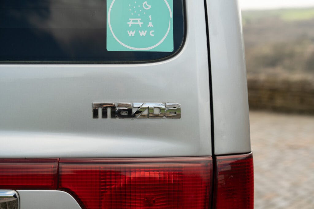 Close-up of the back of a silver 2006 Mazda Bongo Friendee, featuring the Mazda emblem above the right rear taillight. There is a round green sticker on the back window with an illustration of a picnic table, trees, and a moon, and the letters "WWC".