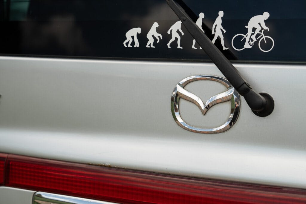 A close-up of the back of a 2006 Mazda Bongo Friendee displays a rear windshield sticker depicting the evolution of humans, ending with a figure riding a bicycle. The Mazda emblem and rear windshield wiper are also visible.