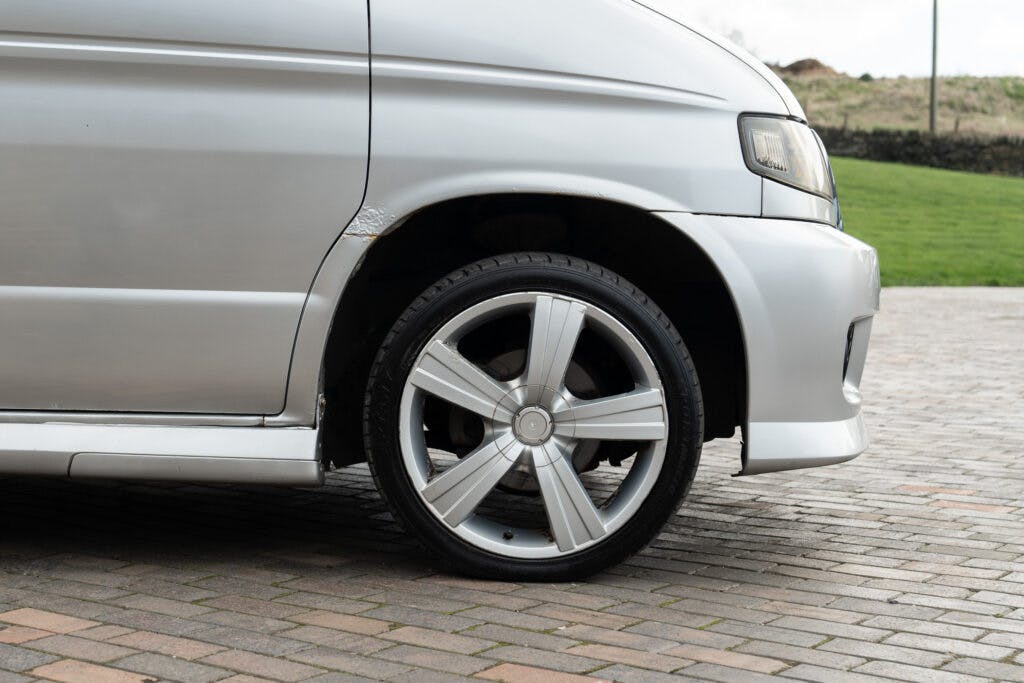 A close-up view of the front passenger side of a silver 2006 Mazda Bongo Friendee. The image focuses on the wheel and tire, which features a silver alloy rim. The car is parked on a brick-paved surface, with a grassy area visible in the background.