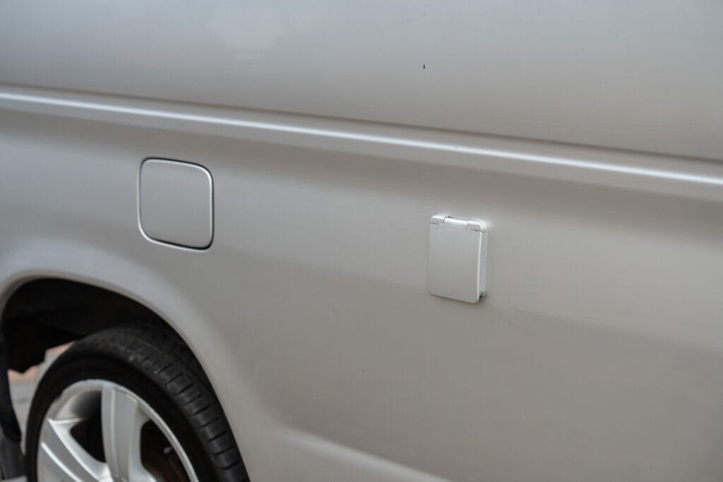 Close-up of the side of a silver 2006 Mazda Bongo Friendee showing the fuel door and a small square compartment cover. The wheel and part of the wheel well are visible in the bottom left corner of the image. The surface of the vehicle appears clean and polished.