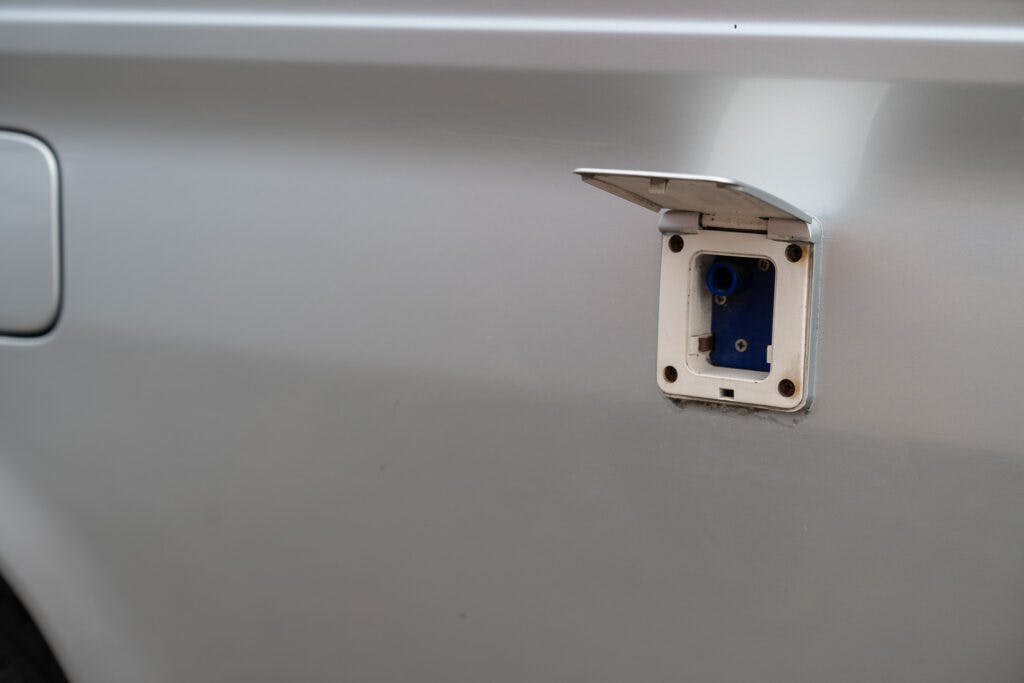 A close-up view of a 2006 Mazda Bongo Friendee's fuel filler area, with the fuel cap removed and the compartment open. The external body of this versatile vehicle is silver, showcasing the circular opening where the fuel nozzle is inserted.