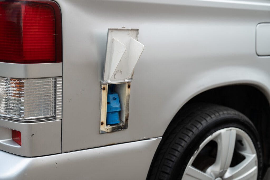 A close-up of a 2006 Mazda Bongo Friendee's rear left side shows an open fuel door exposing a blue propane tank connector. The car's silver exterior and rear light are visible, along with a portion of the rear tire and alloy wheel. The fuel door appears to be made of metal.