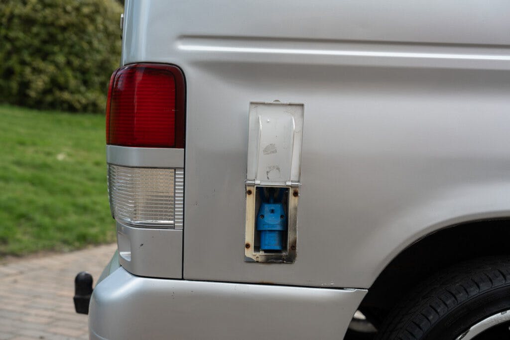 The image shows the rear corner of a 2006 Mazda Bongo Friendee with an open compartment revealing a blue electrical outlet. The vehicle is parked on a paved area with green grass and some bushes visible in the background.