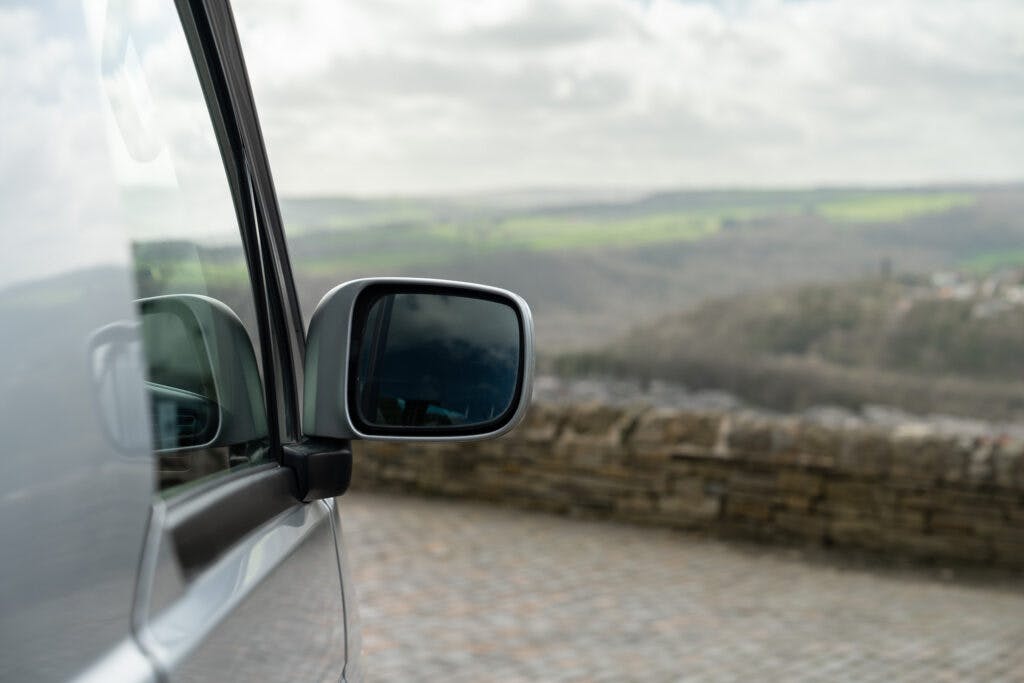 Close-up of the 2006 Mazda Bongo Friendee's silver side mirror reflecting the cloudy sky. The car is parked on a cobblestone driveway, with a stone wall and a scenic, hilly landscape visible in the background.
