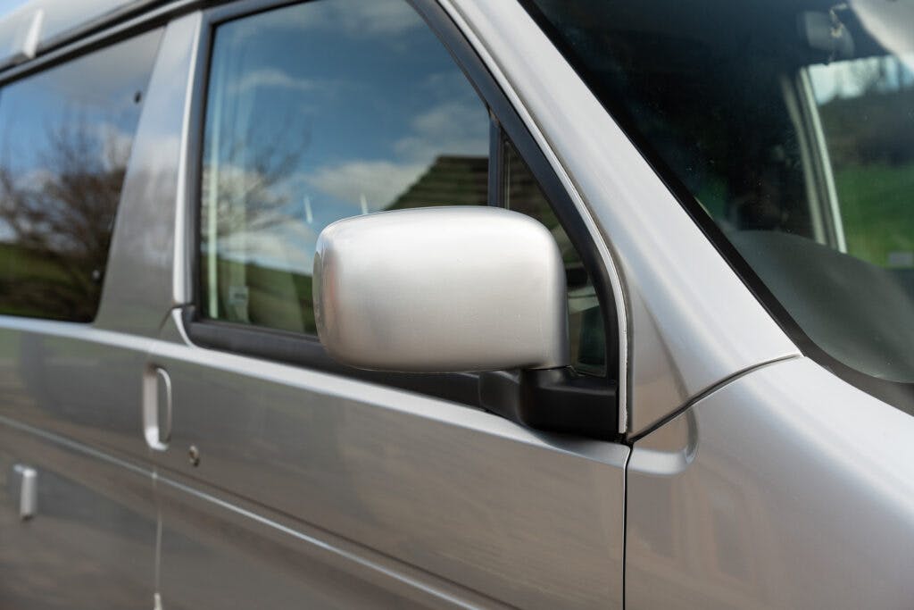 Close-up of a silver side mirror on a 2006 Mazda Bongo Friendee. The van is parked, and in the reflection of the mirror, a house and some trees can be seen. The sky in the background is partly cloudy. The overall setting appears to be a residential area.