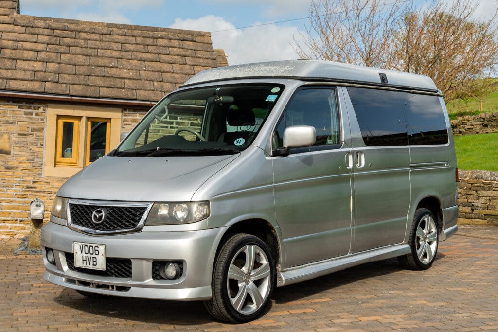 A silver 2006 Mazda Bongo Friendee van is parked on a paved driveway in front of a stone house. The registration plate reads "VO06 HVB". The van has tinted windows and is in good condition under a partially cloudy sky.