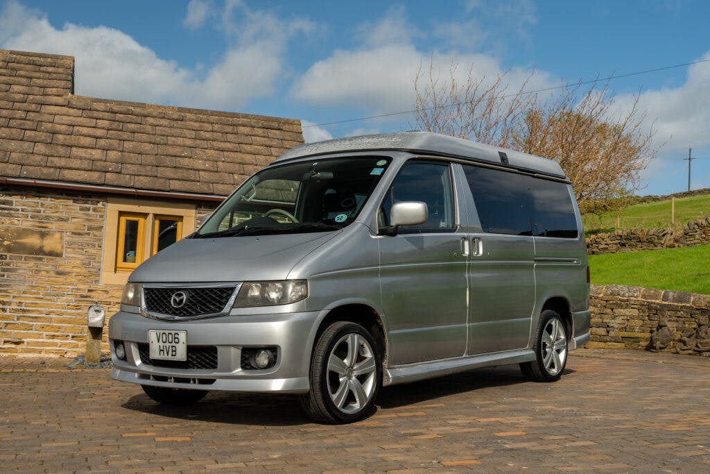 A 2006 Mazda Bongo Friendee camper van is parked on a stone-paved driveway in front of a house with stone walls and a brown tiled roof. The sky is partly cloudy, and there is greenery in the background. The vehicle has a UK license plate reading "VO06 HVB".