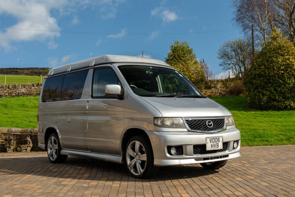 A silver 2006 Mazda Bongo Friendee van is parked on a paved area in an outdoor setting. The van features tinted windows, alloy wheels, and a UK license plate. The background includes a stone wall, green grass, trees, and a blue sky with clouds.