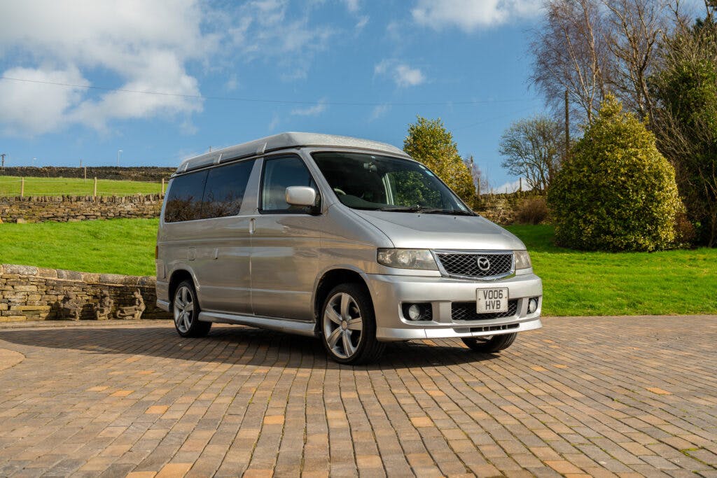 A silver 2006 Mazda Bongo Friendee is parked on a cobblestone surface with a grassy area and trees in the background. The sky is partly cloudy. The license plate reads "VO06 HVB". This van features alloy wheels and a high roofline, perfect for any adventure.