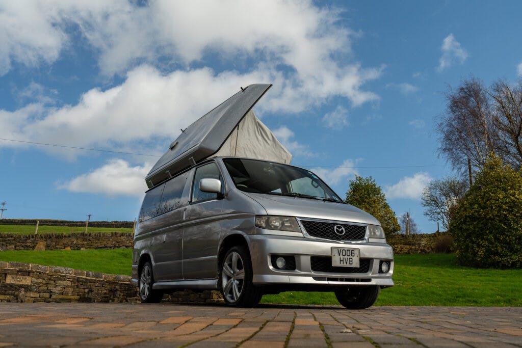 A silver 2006 Mazda Bongo Friendee campervan is parked on a paved surface with its pop-up roof extended. The background features a green lawn, some trees, a stone wall, and a partly cloudy sky. The vehicle has a visible license plate reading "VO06 HVB".