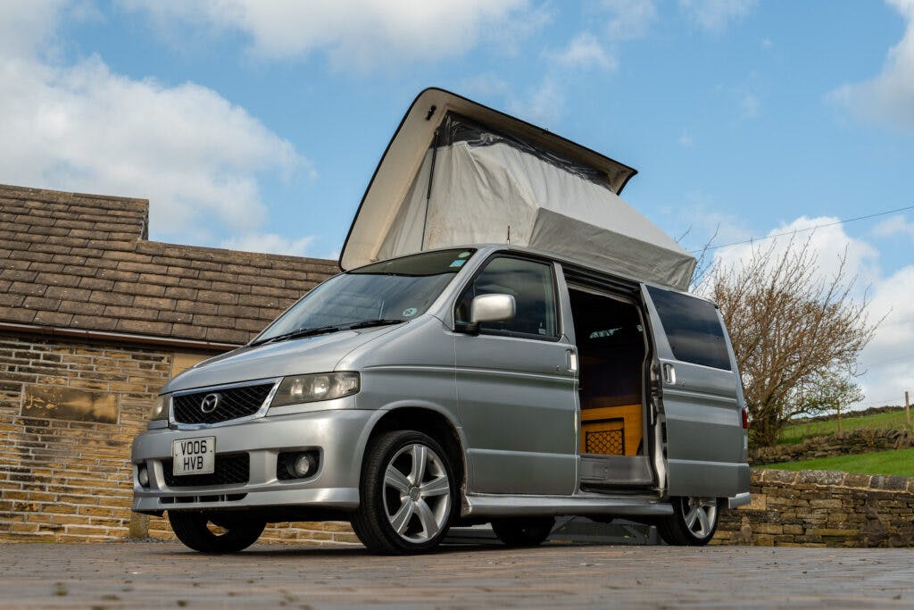 A silver 2006 Mazda Bongo Friendee with an elevated pop-up roof is parked on a brick driveway next to a stone building. The van's side door is open, revealing part of the interior. It is a sunny day with a few clouds in the sky, and trees and grass are in the background.