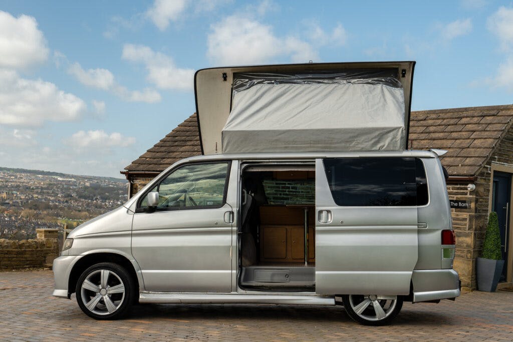 A silver 2006 Mazda Bongo Friendee camper van with its side door open and the pop-up roof extended reveals the interior setup. It is parked on a paved area with a stone building and a scenic landscape in the background under a partly cloudy sky.