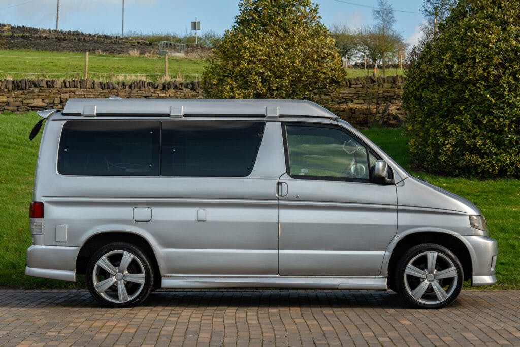 A silver 2006 Mazda Bongo Friendee is parked on a paved area with grass, trees, and a stone wall in the background. The van has tinted windows and six-spoke alloy wheels. The roof is raised, indicating it might be a camper van or a vehicle equipped for longer journeys.