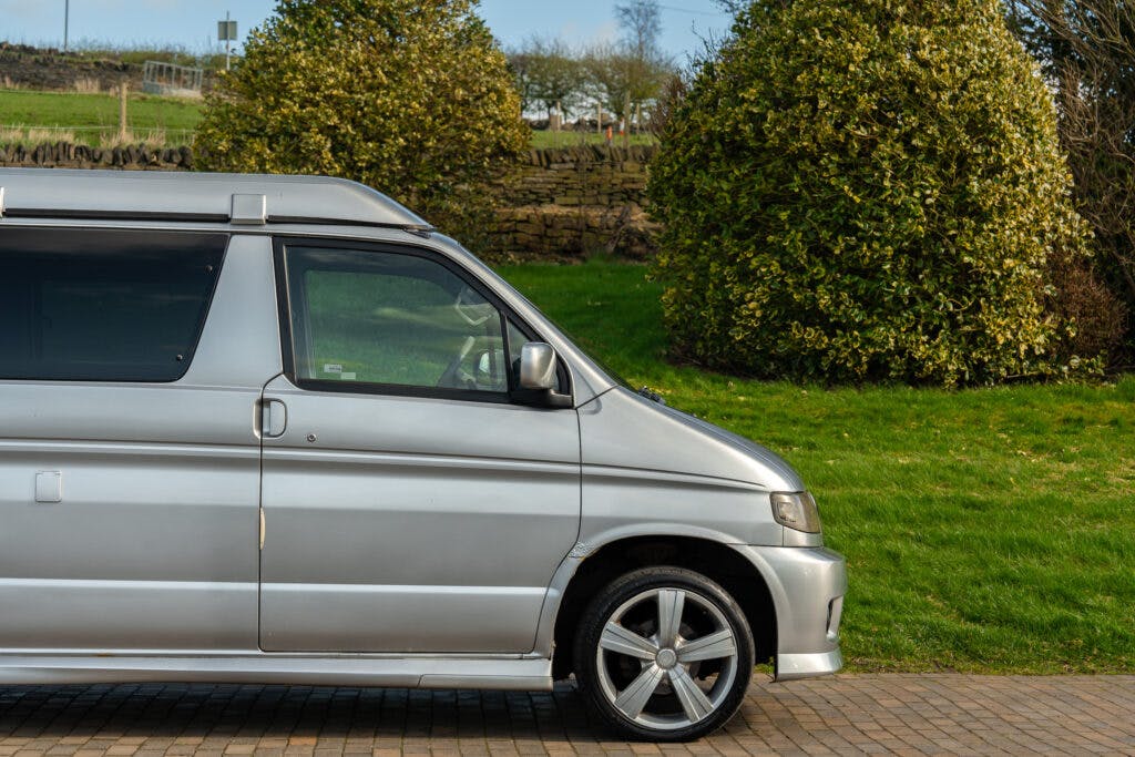 A 2006 Mazda Bongo Friendee, silver in color, is parked on a paved surface beside a grassy area with lush greenery. The front half of the minivan, including the driver's side door and wheel, is visible, with trees and a low stone wall in the background.