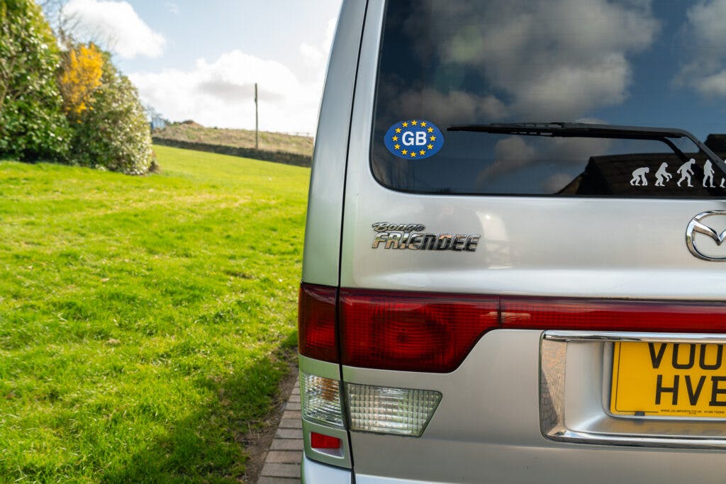 A 2006 Mazda Bongo Friendee, with a silver finish, is parked on a driveway. It features a yellow license plate and a "GB" sticker on the rear window. The surroundings include a green lawn, an orderly hedge, and a partly cloudy sky.