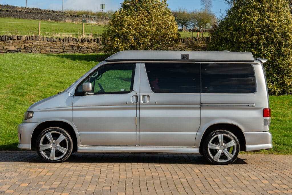 A 2006 Mazda Bongo Friendee is parked on a paved driveway with a grassy hill and stone wall in the background. The silver van boasts tinted windows, a raised roof, and alloy wheels. Trees and shrubs are visible in the surroundings.