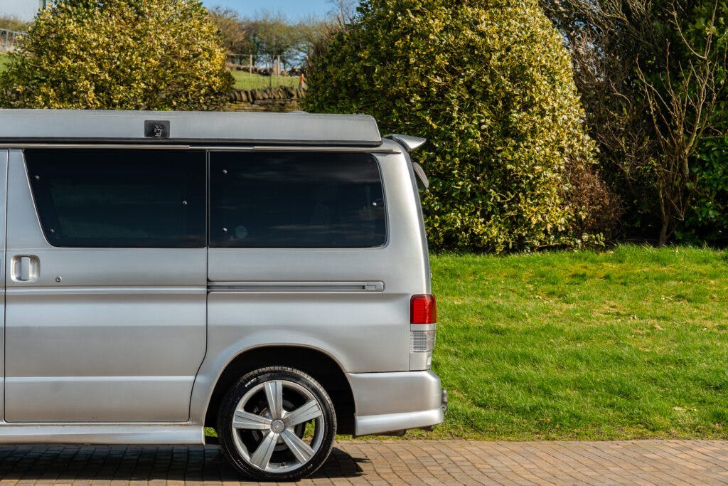 A silver 2006 Mazda Bongo Friendee with tinted windows is parked on a paved surface. The rear half of the vehicle is visible. There is a grassy area with large bushes and trees in the background. The sky is mostly clear.
