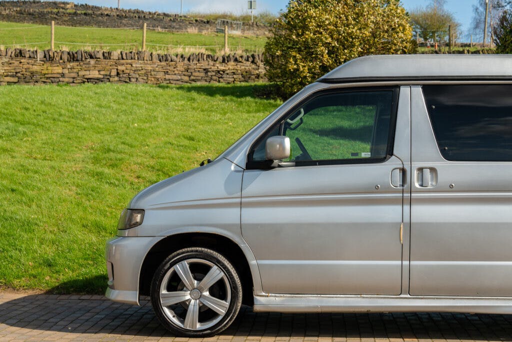 A silver 2006 Mazda Bongo Friendee is parked on a paved driveway beside a grassy area with a stone wall in the background. The image shows the vehicle's front and side, focusing on the driver's door and front wheel.