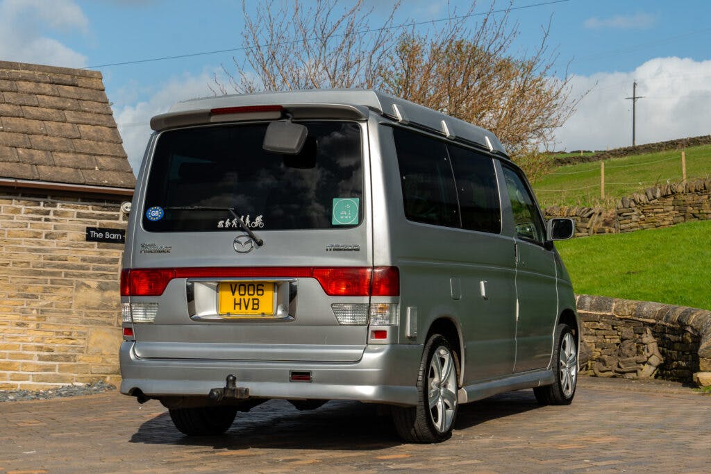 A 2006 Mazda Bongo Friendee, finished in silver, is parked on a paved driveway beside a stone building. The rear of the van is visible, showcasing a yellow license plate with the registration number VO06 HVB. The surrounding area includes some greenery and stone walls.